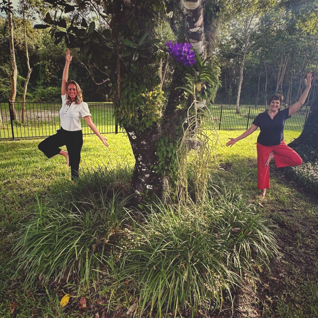 Michelle Natalya Moore on the right side of a tree and a participant on the left side. Both women in a variation of the yoga tree pose.
