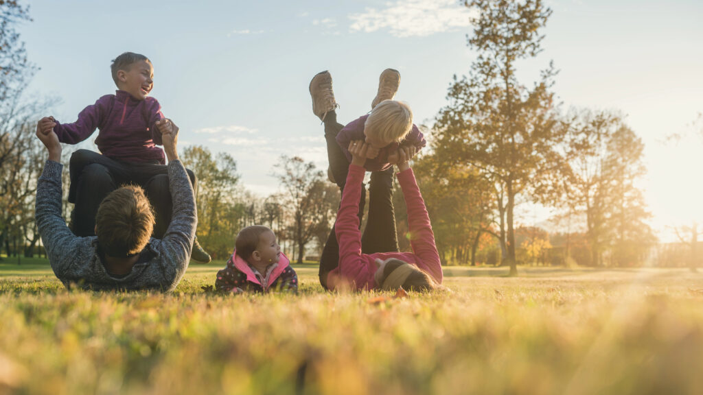 Family with three kids having fun in autumn park—mum and dad lying on the floor each of them lifting one toddler and baby girly lying in grass between them.
