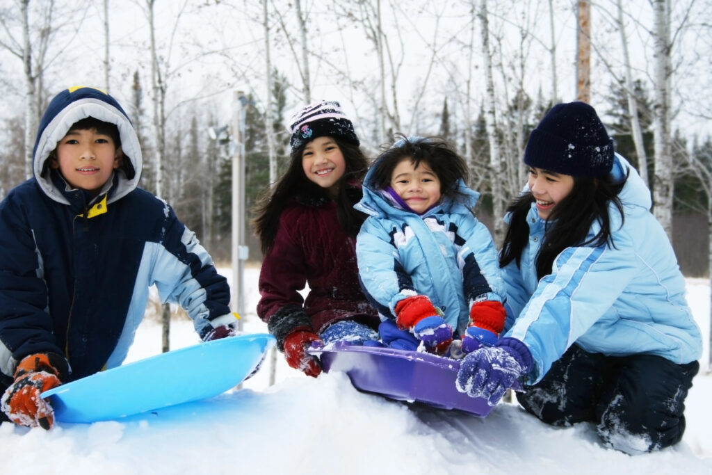Four kids enjoying winter with two sleds at the top of a hill.