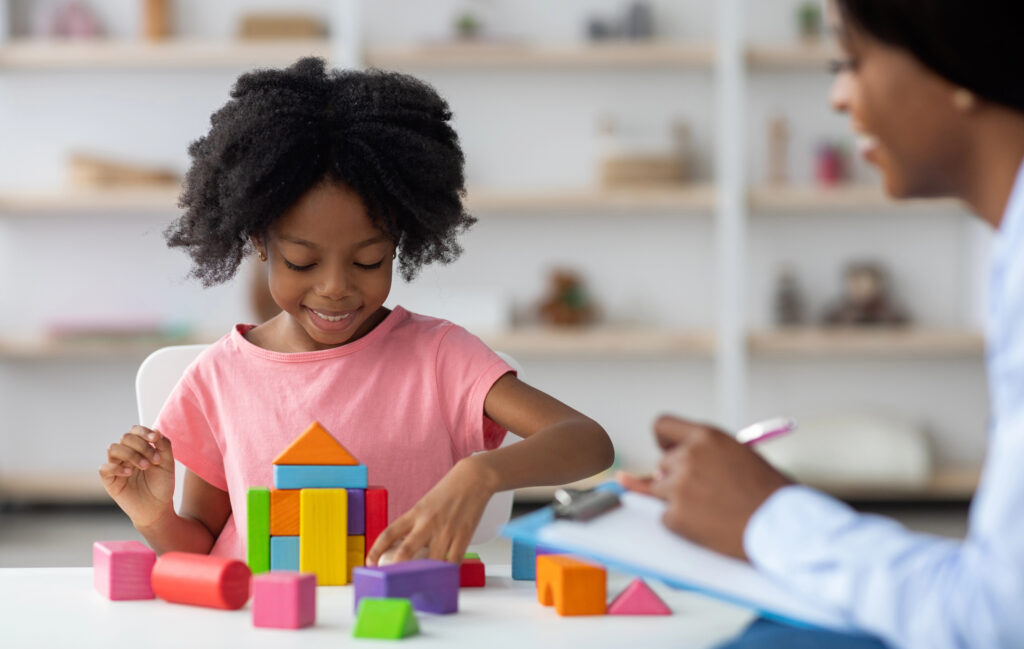 Female child development specialist observing cheerful child preschooler, happy little african american girl sitting at table and making pyramids from colorful wood blocks.