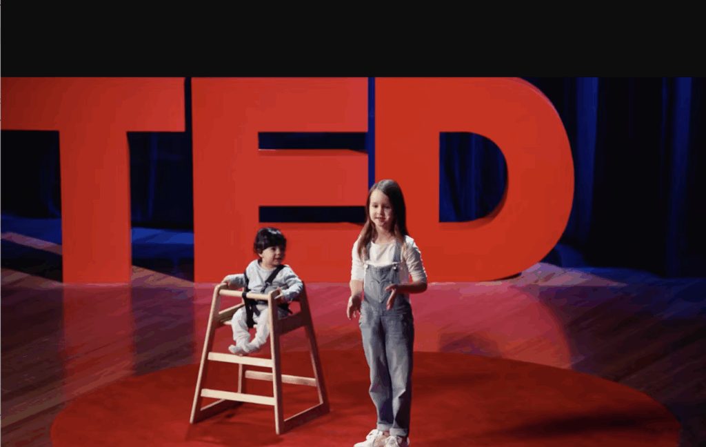 Molly Wright speaking in front of TED Talk sign on stage with young child beside her in a high chair.