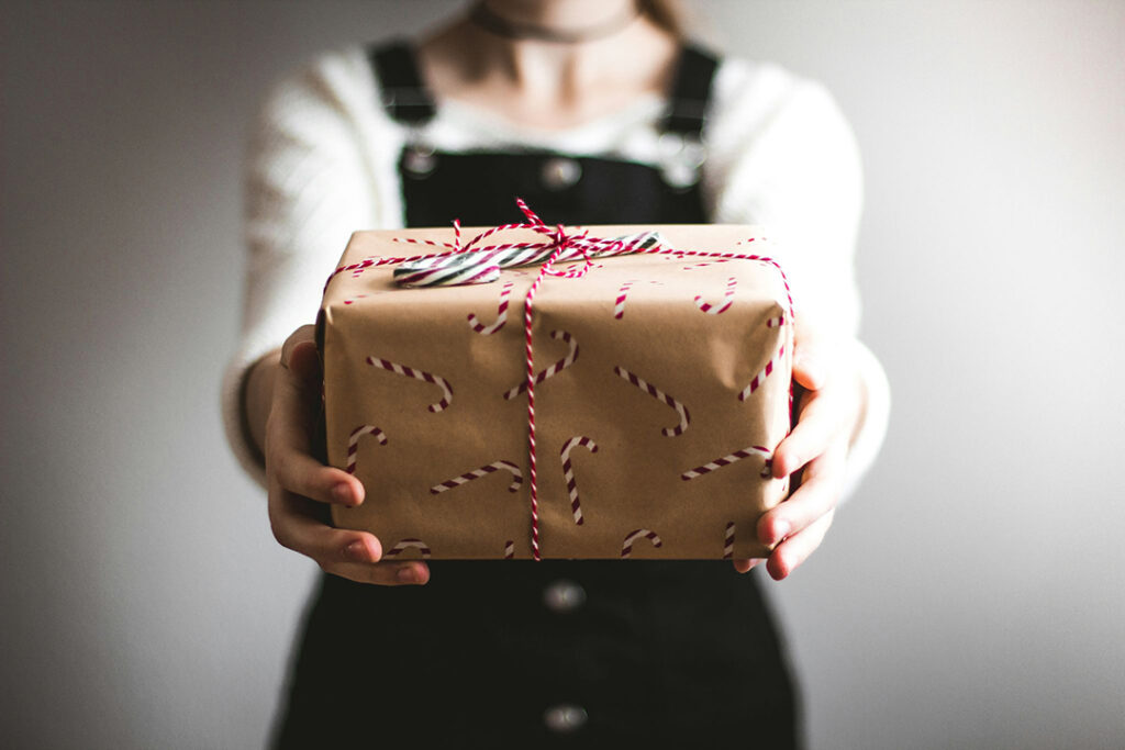teenager in white shirt and black overalls holding a Christmas gift out in front of her that is wrapped in brown paper painted with candy canes, wrapped in a red and white striped string with a candy cane on top.