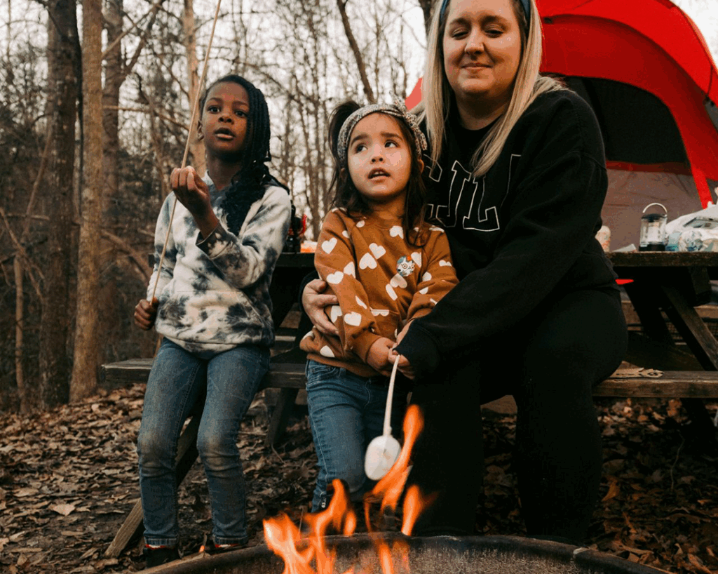 Mother with two daughters sitting on picnic table roasting marshmallows over campfire in bush in front of tent.