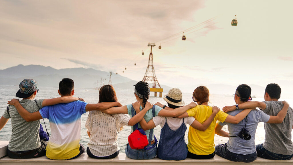 Eight teens sitting on water breakwall with their arms around each others' shoulders looking out at water with gondola and mountain in background.