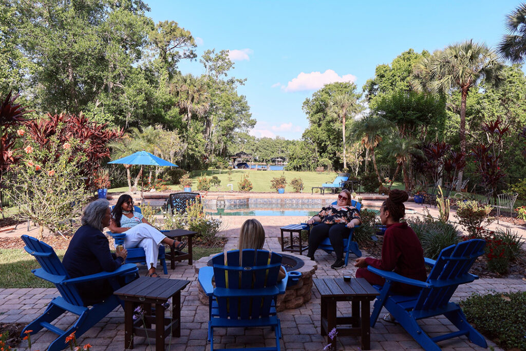 Women sitting on blue adirondack chair around fire pit chatting during the "Women's Retreat: Disconnect to Connect". In the middle ground a pool; background the lake; and all surrounded by trees.